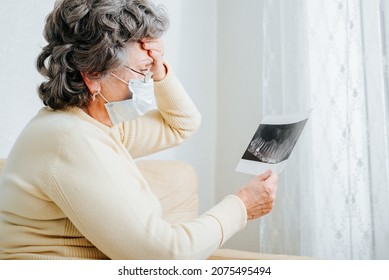 Side View Of Senior Woman Patient In Medical Face Mask Emotionally Analyzing Results Of Medical Examination, Looking At X-ray Of Jaw While Sitting Alone At Home. Seniors And Healthcare, Dentistry.