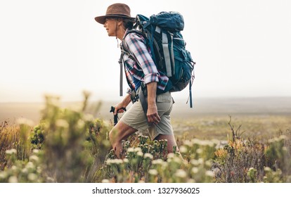 Side View Of A Senior Woman Trekking In The Wild. Woman Wearing Backpack And Hat Walking Through Bushes And Plants During Trekking Holding A Hiking Pole.