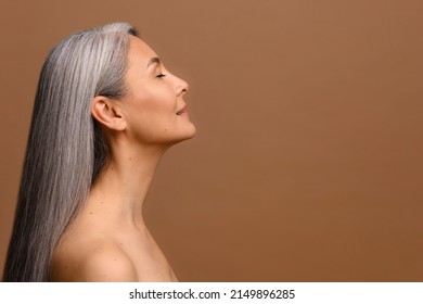 Side View At Sensual Naked Middle-aged Beautiful Asian Woman With Long Grey Hair, Her Eyes Closed, Body And Face Care Concept. Charming Mature Lady Stands In Profile Isolated On Brown Background