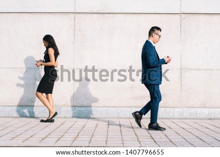 Side view of separate modern multiracial man and woman in formal clothes passing each other busy with smartphones while walking on street 
