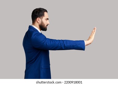Side view of serious bearded man making stop gesture showing palm of hand, conflict prohibition warning about danger, stop bullying, wearing suit. Indoor studio shot isolated on gray background.