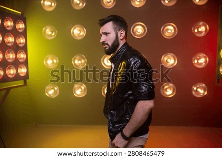 A side view of a serious, bearded man standing with his hands in his pockets. He wears a black leather jacket against a dark background illuminated by round golden lamps and a soft green side glow.