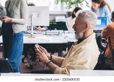 Side View Of Serious Businessman In Casual Clothes Sitting On Chair And Using Mobile Phone While Working On Business Startup With Coworkers In Office