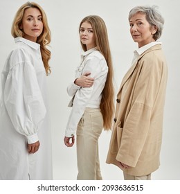 Side View Of Serious Caucasian Family Of Three Female Generations Standing And Looking At Camera. Age And Generation Concept. Grandmother, Mother And Granddaughter. White Background. Studio Shoot