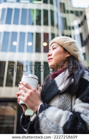 Side view of serious Chinese female in beret and coat standing near modern building with smartphone and thermos with hot beverage in hands