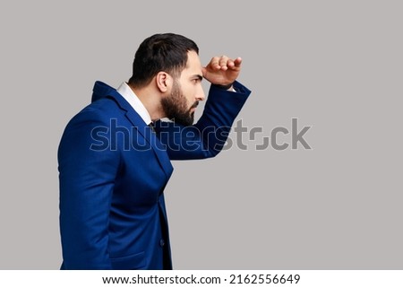 Side view of serious concentrated bearded man watching far away with hand above eyes, looking forward to future, wearing official style suit. Indoor studio shot isolated on gray background.