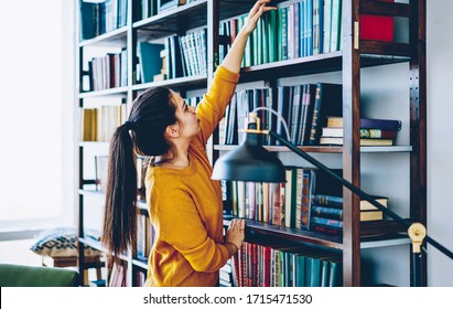 Side view of serious female in casual outfit standing near bookcase and picking book while having rest in library during weekend