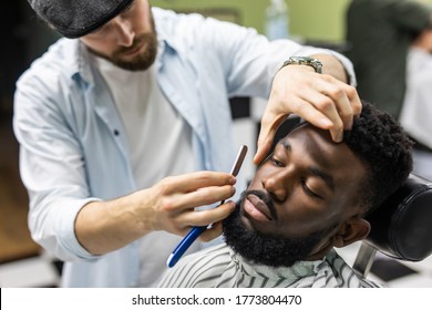 Side view of serious man with stylish modern haircut looking forward in barber shop. Hand of barber keeping straight razor and cutting trendy stripes on head of client. - Powered by Shutterstock