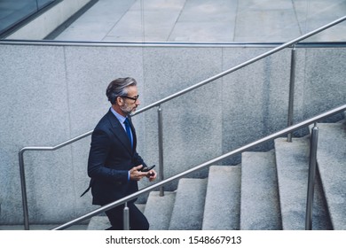 Side View Of Serious Mature Grey-haired Busy Man In Official Suit And Glasses Walking Up Stairs With Digital Modern Smartphone At Street On Urban Background