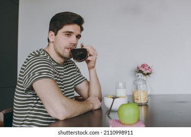 Side View Serious Minded Young Man In T-shirt Eat Breakfast Muesli Cereals With Milk Fruit In Bowl Sit By Table Look Aside Cooking Food In Light Kitchen At Home Alone Healthy Diet Lifestyle Concept.