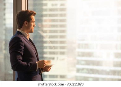 Side view of serious pensive director of prosperous company enjoying cup of coffee in the morning, thoughtful businessman in suit looking through big office window at dawn sunrise city, copy space