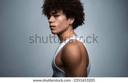 Side view of serious self assured young fit Hispanic male model with curly dark hair in white top gazing at camera against gray background