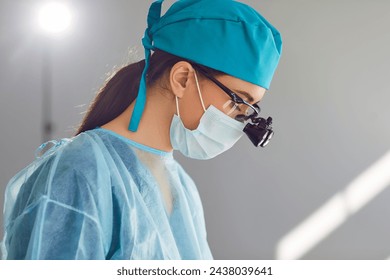 Side view serious skilled woman surgeon wearing face mask, doctor's safety glasses, surgical loupes and blue medical hat doing surgery and saving patient from disease in modern operating theater room - Powered by Shutterstock