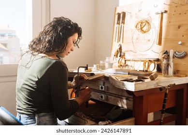 Side view of serious young curly hair female in casual clothes sitting at table and making jewellery while working with equipment in workshop at daylight - Powered by Shutterstock