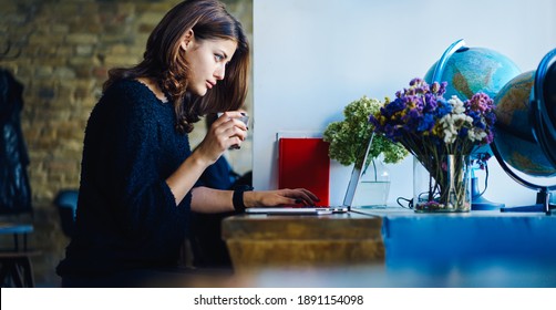 Side View Of Serious Young Remote Worker Using Laptop While Sitting In Cafe At Table With Coffee In Hand And Flower And Globe Near Computer