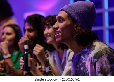 Side view of several young intercultural friends watching broadcast of match at gathering with focus on happy guy wearing beanie hat - Powered by Shutterstock