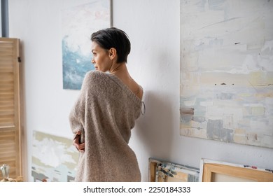 Side view of sexy brunette artist in sweater standing near paintings in studio - Shutterstock ID 2250184333