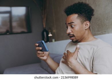 Side View Shocked Disapponted Scared Sad Young African American Man Wearing Beige T-shirt Sit On Grey Sofa Indoor Apartment Holding Mobile Cell Phone Surfing Internet Rest On Weekends Stay At Home.