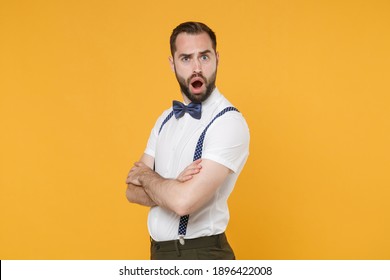Side view of shocked perplexed young bearded man 20s wearing white shirt bow-tie suspender posing standing holding hands crossed looking camera isolated on yellow color background studio portrait