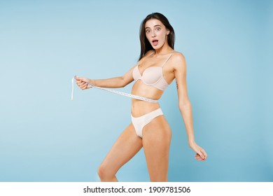 Side view of shocked worried young brunette woman 20s wearing beige brassiere underwear standing posing measuring waist with measure tape isolated on pastel blue colour background studio portrait