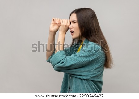 Side view of shocked young woman standing with hands on eye monocular gesture and looking away with surprised face, wearing casual style jacket. Indoor studio shot isolated on gray background.