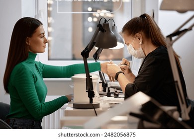 Side view shot of female nail tech wearing protective mask while doing manicure for young brunette woman during pampering beauty treatment in nail salon