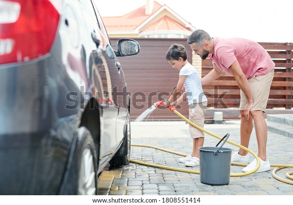 Side view shot of happy father\
watching his little son washing car wheel with water, copy\
space