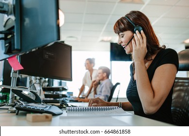 Side View Shot Of Happy Mature Business Woman Talking On Headset While Working At Her Desk With People Working In Background.