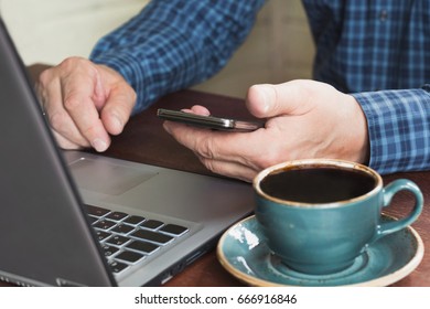 Side view shot of a man's hands using smart phone and laptop sitting at wooden table with cup of black coffee in interior.  Close up.
