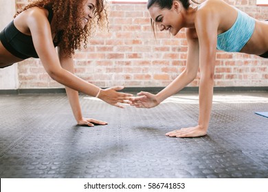 Side View Shot Of Two Young Women Exercising In Gym With Their Hands Touching. Female Friends Doing Pushups Together.