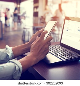 Side View Shot Of Young Business Man Working On His Laptop And Using Smart Phone Sitting At Wooden Table In A Coffee Shop With Retro Filter Effect