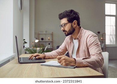 Side view shot of young man wearing glasses using laptop. Businessman sitting at desk looking at computer screen and writing down list to do in notepad in comfortable office