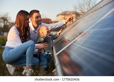 Seitenansicht einer jungen modernen Familie mit einem kleinen Sohn, der an einem sonnigen Tag mit Solarpaneelen vertraut wird, grünes Alternativenergiekonzept
