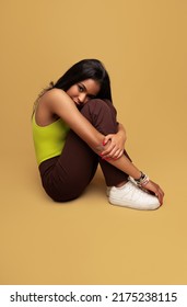 Side View Of Shy Teen Model In Trendy Clothes With Black Hair Embracing Knees And Looking And Camera While Sitting Against Ocher Background