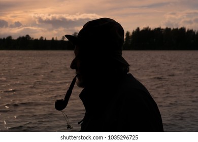Side view silhouette portrait of an old fisherman in rain hat and coat, standing by the ocean smoking a pipe in the pink evening sunlight