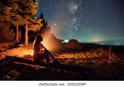 Side view of silhouette of woman relaxing near forest in campsite. Female sitting near bonfire and admiring starry sky. Concept of adventure and night camping in the mountains.
