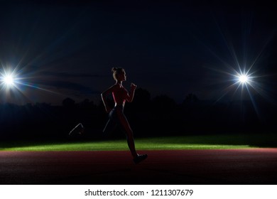 Side View Of Silhouette Of Woman Running Forward In Darkness On Stadium Only With Two Lights From Two Sides. Sports Girl Training At Night Alone, Doing Big Steps To Competition. Concept Of Sport.