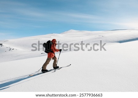 Side view of skier in orange jacket moving up gentle snowy mountain slope on skis