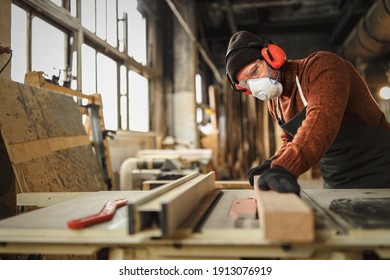 Side view of skilled carpenter in protective respirator and headphones cutting lumber plank on workbench in professional workshop