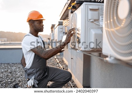 Side view of skilled man typing on wireless laptop while kneeling near hanging air conditioner outdoors. African american specialist using modern technologies for quickly repairing of cooling system.
