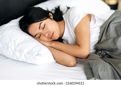 Side view of a sleeping asian girl in a cozy bed in apartment, pleasant dreams, healthy sleep. Pretty chinese woman in white t-shirt under the blanket in bed in the morning, sees sweet dreams - Shutterstock ID 2160194511