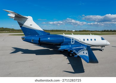 Side view, slightly high angle, full photograph of a modern blue and white private executive jet parked on an airport tarmac under a blue sky with light clouds.