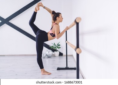 Side view of slim ballerina holding on to barre while raising leg high and stretching body muscles in light studio