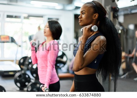 Side view of slim black african-american fitness instructor and asian lovely woman doing fitness exercise work with dumbbells together in the gym