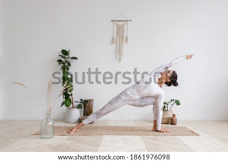 Side view of slim pretty positive young brunette woman doing Utthita parsvakonasana exercise, Extended Side Angle pose, on mat on floor surrounded by houseplants on white wall. Yoga and pilates