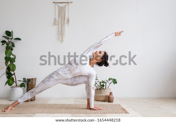 Side view of slim pretty positive young brunette\
woman doing Utthita parsvakonasana exercise, Extended Side Angle\
pose, on mat on floor surrounded by houseplants on white wall. Yoga\
and pilates