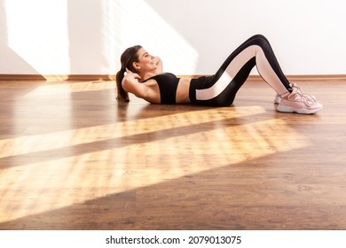 Side view of slim sportive woman doing crunch exercise, training abdominal muscles, wearing black sports top and tights. Full length studio shot illuminated by sunlight from window. - Shutterstock ID 2079013075