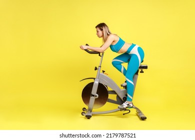 Side view of slim woman with perfect body riding exercise bike at home, making an effort to lose a few extra pounds, wearing blue sportswear. Indoor studio shot isolated on yellow background. - Powered by Shutterstock