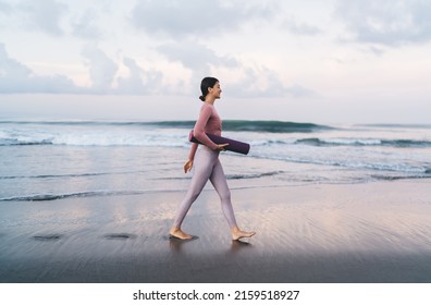 Side view of slim woman in sportive clothes holding mat and walking near calm sea waves spending weekend morning for training at nature, charming female in tracksuit choosing place for stretching - Powered by Shutterstock