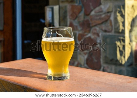 A side view of a small sample glass of pale ale craft beer. The liquid has a yellow tint. A clear beer glass sits on the edge of a wooden table at a microbrewery. The background has a rock wall.